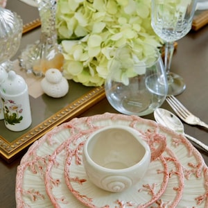 May include: A ceramic dinnerware set featuring a white plate, a smaller white saucer, and a white cup, all decorated with raised, textured pink coral-like patterns along the edges. The cup has a distinct spiral shell design on its exterior.