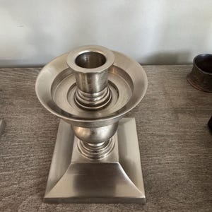May include: A silver-colored candlestick holder with a square base and a round top. The candlestick has a classic design, with a flared base and a cup-shaped holder for the candle. The metal has a brushed finish.
