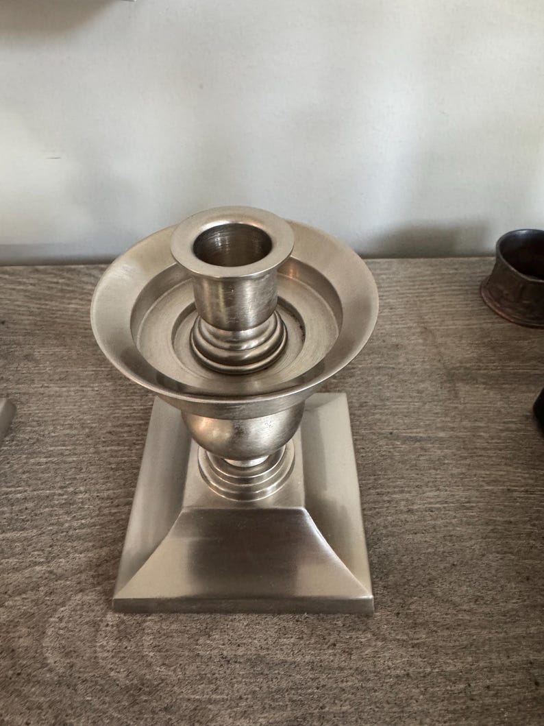 May include: A silver-colored candlestick holder with a square base and a round top. The candlestick has a classic design, with a flared base and a cup-shaped holder for the candle. The metal has a brushed finish.