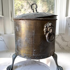 May include: Antique brass container with a lid and a handle. The container has a lion's head ring pull and rests on three claw feet. The brass has a weathered patina, giving it a vintage appearance. The container is cylindrical in shape.