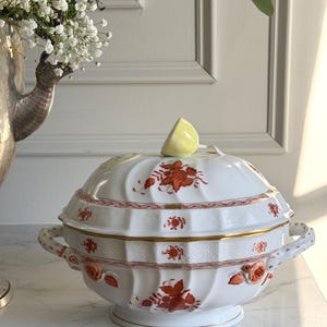 May include: A white porcelain soup tureen with a lid, adorned with red floral patterns and gold trim. The tureen has two handles and a base. A yellow lemon slice rests on the lid. The tureen is on a marble surface.