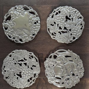 May include: Four silver-colored metal coasters with intricate grape and leaf designs. Each coaster is round, with detailed relief patterns. One coaster has a smooth center, while the others feature grape clusters and foliage.