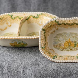 May include: A cream-colored ceramic divided serving dish and a matching rectangular bowl, both featuring a decorative yellow and green border pattern and a central landscape illustration depicting a building and trees in muted tones with a rope-like rim.