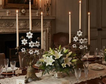May include: A dining table set with two ornate metal candelabras featuring white flower accents and tall taper candles, a central silver bowl filled with white lilies and greenery, crystal glassware, and porcelain plates with floral motifs.