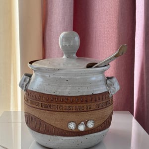 May include: A ceramic lidded jar with a spoon. The jar is off-white with a brown band featuring the text "ALL THINGS THROUGH CHRIST WHO STRENGTHENS ME." The jar has a handle on the lid and two side handles. Three white flower-like designs are on the brown band.