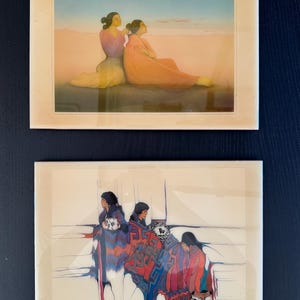 May include: Two framed prints of Native American art. The top print shows two women sitting on a sandy beach, one is braiding the other's hair. The bottom print shows four women sitting in a row, wearing colorful blankets.