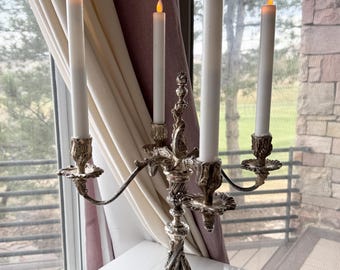 May include: Silver candelabra with four candle holders and ornate details. The candelabra is holding four white candles with a warm glow.