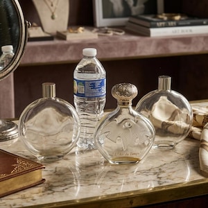 May include: Two clear, round glass perfume bottles with silver caps and a central glass perfume bottle featuring a decorative silver stopper and embossed bird design with the text L'Air du Temps, positioned near a plastic water bottle.