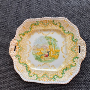 May include: A square white ceramic serving plate with a green and gold floral design. The center of the plate features a painted scene of a couple in a garden setting.