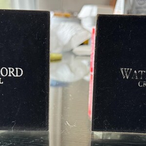 May include: Two black Waterford Crystal boxes are displayed. The boxes are square and have the Waterford logo and the word "CRYSTAL" printed in white. The boxes are sitting on a clear surface.