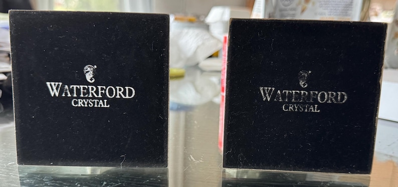 May include: Two black Waterford Crystal boxes are displayed. The boxes are square and have the Waterford logo and the word "CRYSTAL" printed in white. The boxes are sitting on a clear surface.