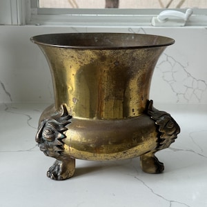 May include: A vintage brass vase with a flared top and three decorative feet. The vase has a tarnished, aged appearance, with intricate details on the feet, which are sculpted with a face design. The vase is likely used for decorative purposes.