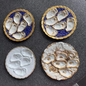 May include: Four round ceramic oyster plates, each featuring six molded indentations for serving. Two plates have deep blue backgrounds with gold floral patterns and gold rims, one is plain white with a gold rim, and one has a white and gold floral design.