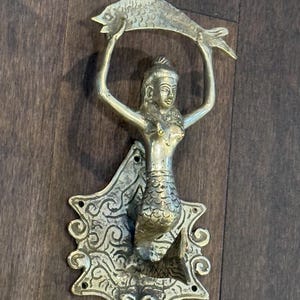 May include: A brass door knocker in the shape of a mermaid holding a fish. The mermaid is detailed with scales and has a crown on her head. The knocker is mounted on a decorative plaque with intricate designs.