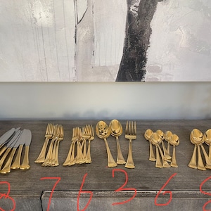 May include: A set of gold-toned flatware, including knives, forks, and spoons, arranged in rows on a wooden surface. The flatware is arranged in groups of 8, 7, 6, 3, 6, and 8.