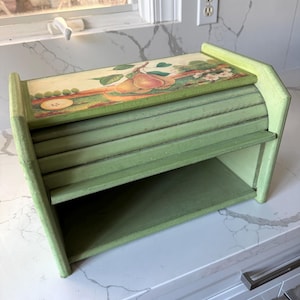 May include: A green wooden bread box with a roll-top lid. The lid features a painted scene of pears and flowers. The box has a rectangular shape with a shelf inside. The bread box is painted in shades of green.