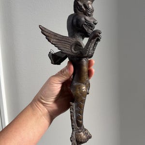 May include: A dark bronze-toned metal sculpture featuring a winged creature with a feline head, a crest-like mane, and a slender, elongated body that tapers down to a decorative base. The figure has detailed wings and a textured surface finish.