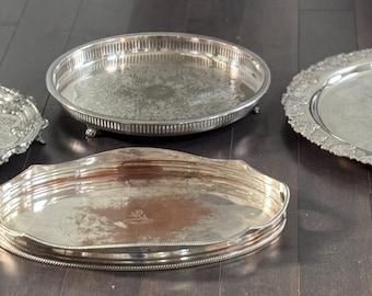 May include: Four silver serving trays of various shapes and sizes are displayed on a dark wood surface. The trays include round, oval, and rectangular designs, each with unique decorative edges and patterns. The trays are reflective and appear to be antique.