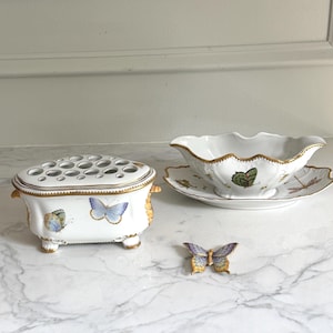May include: A white porcelain flower frog, gravy boat with saucer, and a butterfly figurine. The flower frog has a lid with multiple holes and butterfly decals. The gravy boat and saucer have gold trim and butterfly and dragonfly designs. The butterfly figurine is purple and gold.