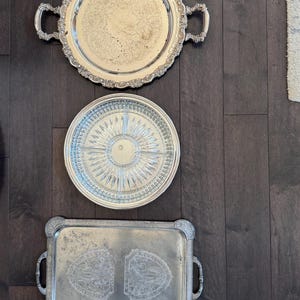 May include: Three silver serving trays of varying shapes and designs are displayed on a dark wood surface. The trays include a round tray with handles, a divided tray, and a rectangular tray with handles.
