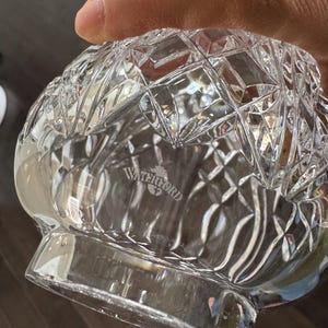 May include: Clear crystal glass bowl with a diamond-cut pattern. The bowl has a rounded base and a flared rim. The word "WATERFORD" is etched on the bottom of the bowl. The bowl is held by a person.