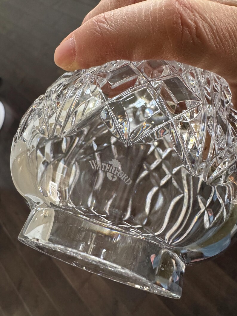 May include: Clear crystal glass bowl with a diamond-cut pattern. The bowl has a rounded base and a flared rim. The word "WATERFORD" is etched on the bottom of the bowl. The bowl is held by a person.
