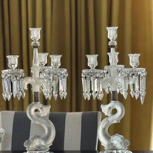 May include: A pair of clear crystal candelabras featuring a central dolphin-shaped base with frosted detailing. Each candelabra has five candle holders arranged in a tiered design, adorned with hanging crystal prisms that catch the light.