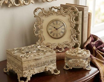 May include: A rectangular cream-colored decorative box with ornate filigree, a matching round glass trinket jar with a lid, and a square analog clock featuring Roman numerals, all finished in a distressed cream tone with intricate scrollwork.