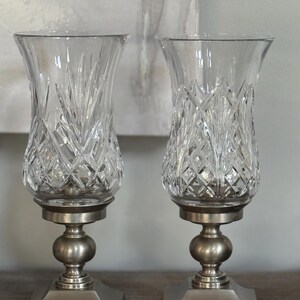 May include: Two clear glass candle holders with diamond and leaf-like cut patterns. Each holder sits on a silver-colored metal base with a square bottom, a round center, and a circular top. The candle holders are displayed on a wooden surface.