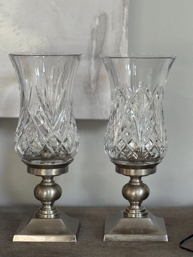 May include: Two clear glass candle holders with diamond and leaf-like cut patterns. Each holder sits on a silver-colored metal base with a square bottom, a round center, and a circular top. The candle holders are displayed on a wooden surface.