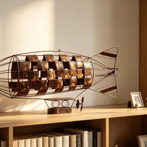 May include: A decorative metal airship sculpture featuring a wireframe structure with a central section composed of overlapping, weathered copper-toned rectangular plates. The model includes a small gondola base with a propeller and tail fins.
