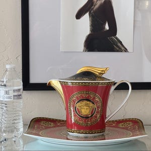 May include: A red and gold ceramic tea set with a Medusa head emblem. The set includes a teapot with a gold handle and lid, and a matching rectangular tray. The teapot and tray are on a glass surface.