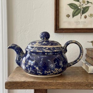 May include: A ceramic teapot featuring a deep blue glaze with a white mottled or sponge-painted pattern. The teapot has a rounded body, a curved spout, a matching lid with a small knob handle, and a C-shaped handle on the side for pouring.