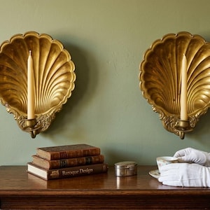May include: Two ornate, gold-colored wall sconces shaped like scallop shells, each holding a cream-colored candle. Below, a wooden table displays antique books and a small silver box. The background is a muted green.