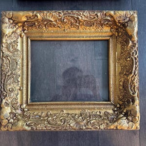 May include: A rectangular picture frame with a metallic gold finish, featuring an ornate, raised relief pattern of scrolling foliage and floral motifs along the wide border, surrounding a central rectangular opening with a dark interior.