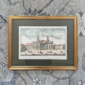 May include: Framed architectural print featuring a detailed illustration of a grand building with multiple stories, red-roofed towers, and a bustling courtyard. The print is set within a gold-colored frame and a dark green mat.