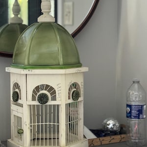 May include: A decorative birdcage with a white distressed finish. The cage features arched windows, metal bars, and green accents. The top is a green dome with a white finial. A mirror and water bottle are in the background.