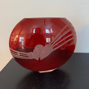 May include: A round, red glass vase with a heart and ray design. The vase has a smooth, glossy finish and a clear base. The design is etched into the glass, creating a frosted effect. The vase is likely used for decorative purposes.