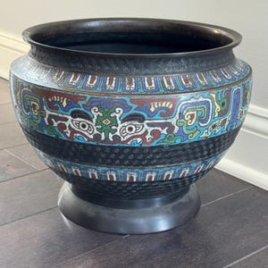 May include: A decorative, antique bronze planter with intricate enamel detailing. The planter features a dark bronze rim and base, with a central band of black geometric patterns. The body is adorned with colorful floral and geometric designs in blue, red, and green.