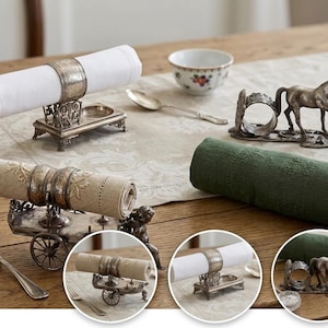 May include: A collection of silver-toned metal napkin holders featuring three distinct designs: a rectangular base with ornate scrollwork, a cart-shaped holder with wheels, and a sculptural horse figure. Each piece holds a rolled fabric napkin.
