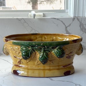 May include: A yellow ceramic planter with a green rim and decorative green artichoke-like designs. The planter has a wide, oval shape with handles on each side and a pedestal base. The edges show a distressed, aged appearance.