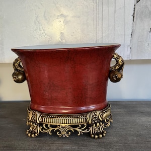 May include: A red ceramic planter with gold accents and a decorative base. The planter has a white interior and a textured exterior.