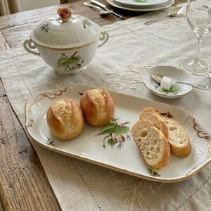 May include: A white porcelain serving tray with gold trim and a botanical berry illustration, holding two whole bread rolls and two sliced baguette pieces. The tray features a scalloped edge and a matching lidded tureen with a rose finial.