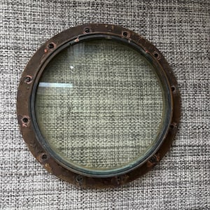 May include: A round, brass porthole with a glass window. The porthole is set against a textured, gray background.