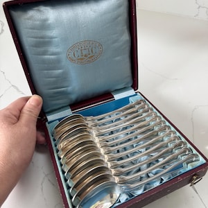 May include: A set of silver spoons in a burgundy box with a light blue satin lining. The spoons are arranged neatly, reflecting light. The box has a gold emblem that reads "Christofle & Cie, Manufacture d'Orfevrerie, Paris."