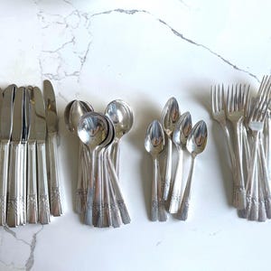 May include: A collection of vintage silverware, including knives, spoons, and forks, arranged on a white marble surface. The silverware has a shiny, reflective surface and a decorative floral pattern on the handles. The utensils are grouped together in sets.
