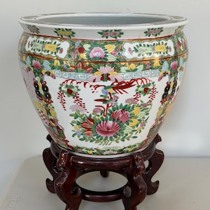 May include: A large, round, white ceramic fishbowl planter featuring intricate, colorful hand-painted floral patterns, butterflies, and birds in shades of pink, green, yellow, and red. The bowl rests on a dark brown, carved wooden display stand.