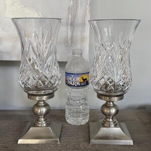 May include: Two clear glass candle holders with a diamond pattern, set on a silver-colored base. The holders have a flared top and a square base. A bottle of water is between them.