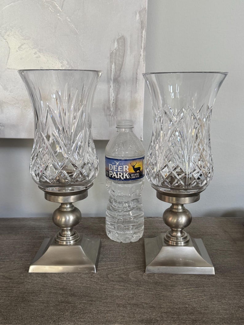 May include: Two clear glass candle holders with a diamond pattern, set on a silver-colored base. The holders have a flared top and a square base. A bottle of water is between them.