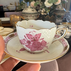 May include: White teacup and saucer set with a pink floral design and gold trim. The teacup has a textured surface and a scalloped rim. The saucer has a matching floral pattern and gold detailing. A classic tea set.
